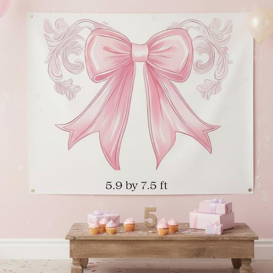 Large pink bow wall decoration with dimensions on a light pink wall, below which is a wooden table with cupcakes and a cake.