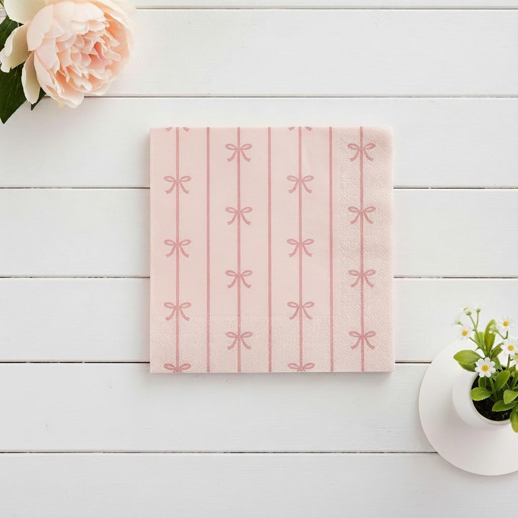Pink dessert napkin with bow pattern on a white wooden surface with flowers and a plant.