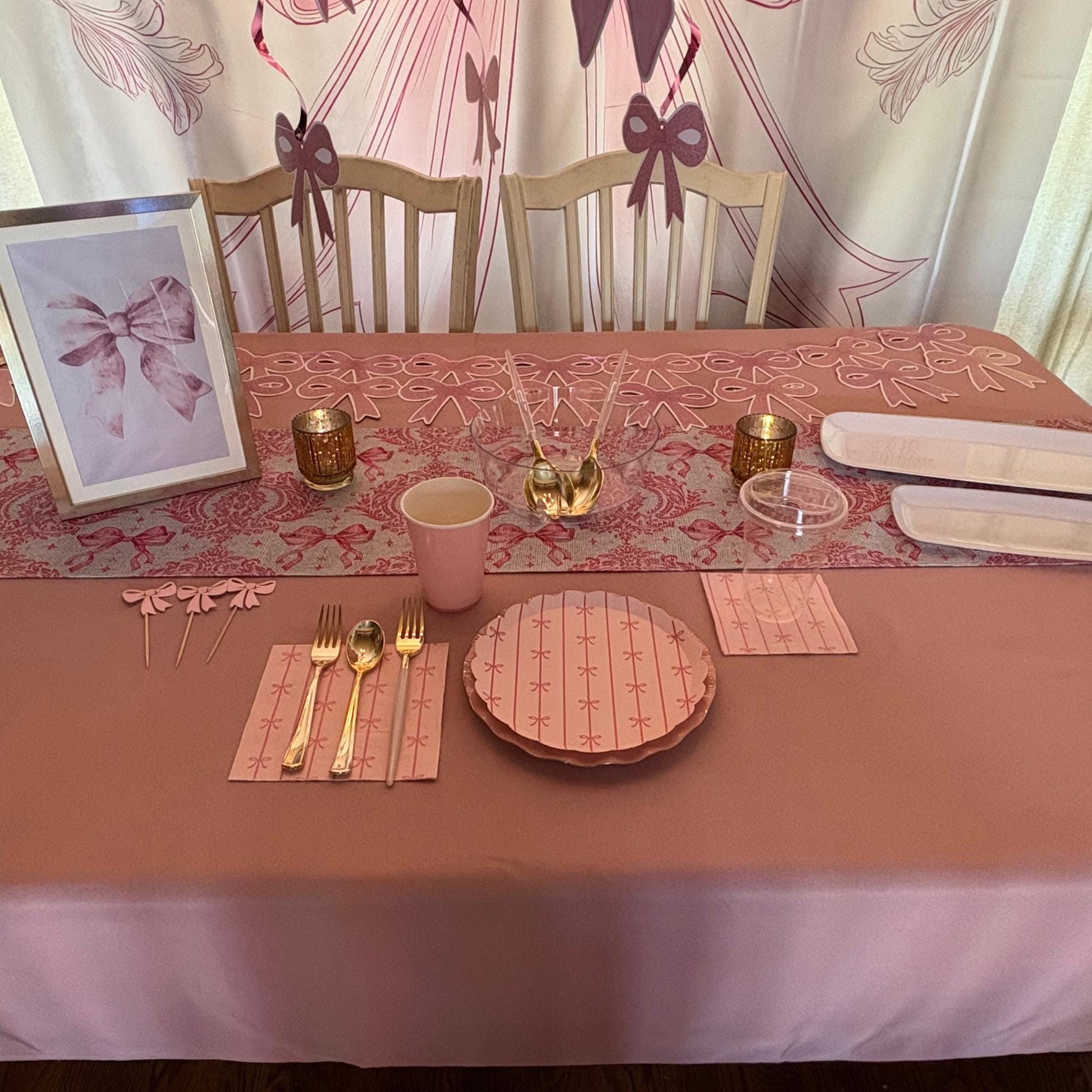 Dining table set with pink tablecloth, plates, cups, and cutlery against a decorative curtain background.